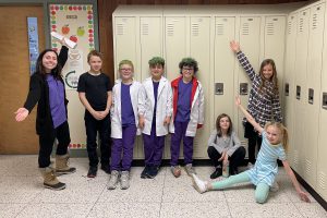 a group of students in costume pose with their teacher in front of a row of school lockers in a hallway