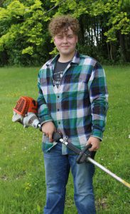 a young man wearing a plaid shirt and jeans holds a weed whacker
