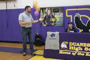 a man with a beard speaks to a young woman sitting at a table in a gymnasium