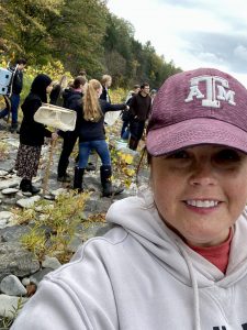 a teacher wearing a red hat takes a selfie with students behind her doing an experiment in the creek