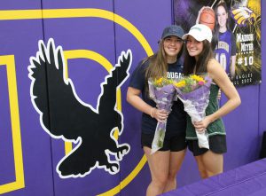 two young women wearing ball caps hold flowers and stand in front of a purple wall in a gymnasium