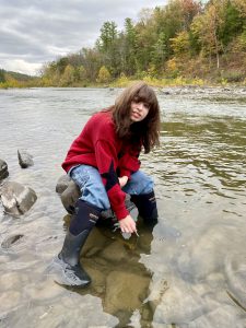 a young teen girl wearing a red coat sits on a rock in the middle of a creek 