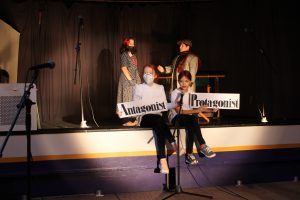 two young girls wearing white shirts and black pants sit on the edge of a stage and hold up signs saying "antagonist" and "protagonist."