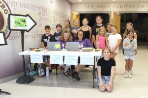 two adult women wearing purple shirts sit behind a table surrounded by young student-actors