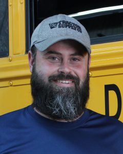 a man with a beard stands next to a school bus