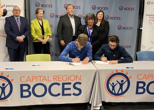 two male students sign a piece of paper as adult dignitaries look on