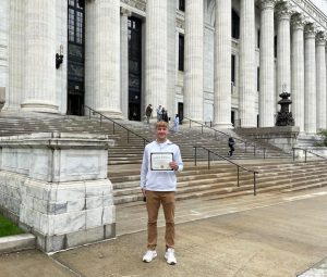 a young man stands in front of a grand staircase, while holding a certificate