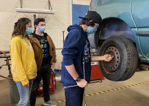 a young male student works on a car's tire, while two other students look on