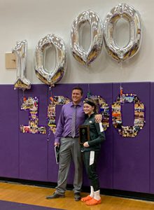 a young female athlete poses with her coach, in front of a sign commemorating her 1,000th career basketball point.