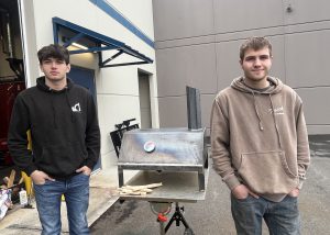 two male teenagers stand outside in a parking lot next to a grill