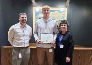 a tall young man stands in between an adult male and an adult female, while holding a certificate