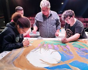 students paint scenery, with instruction from their teacher