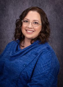 a woman with dark hair and glasses, wearing a blue top, smiles