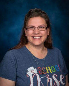 a woman wearing glasses and a t-shirt that says school nurse, smiles at the camera