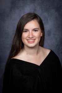 a young woman with dark hair, wearing a black v neck top poses in front of a backdrop