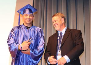 a young man wearing a blue graduation cap and gown stands next to a smiling man on a stage 