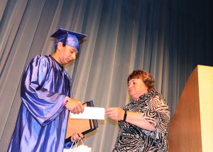 a woman presents an award to a teenager wearing a blue graduation cap and gown on a stage. 