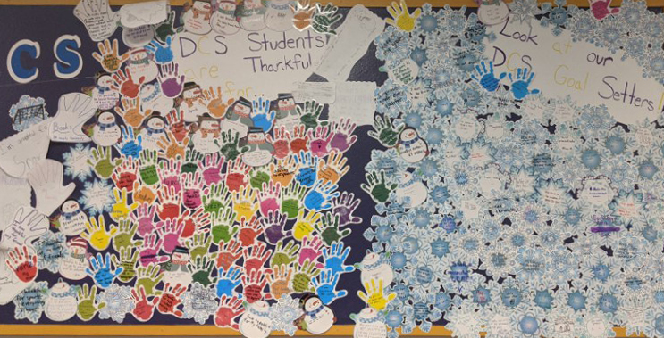 a wideframe view of a collage on a bulletin board showing paper cutouts of handprints and snowflakes showing handwritten notes from students sharing what they are thankful for and what their goals are