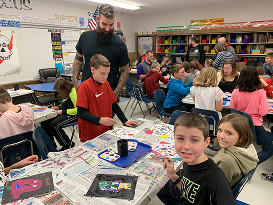 girls and boys seated at tables, painting, as Chad Foster looks on