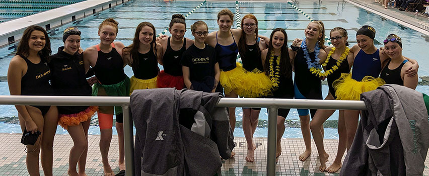 swim team standing in front of school, some wearing tutus