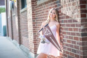 a girl wearing a pink dress leans against a brick wall and holds a banner that reads "St. Boneventure."