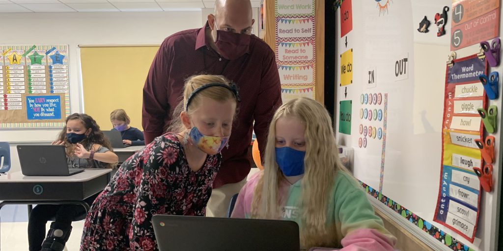 two elementary students wearing masks work together before a laptop while a teacher wearing a mask looks over their shoulders
